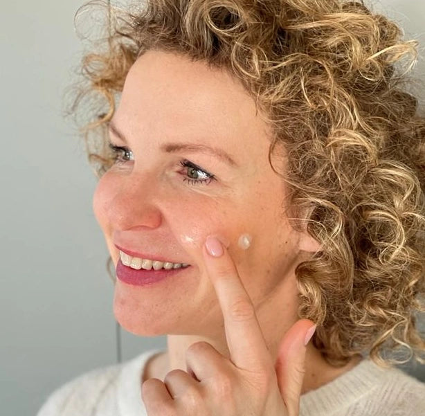 Lachende Frau mit natürlicher, sensibler Gesichtshaut trägt Creme auf